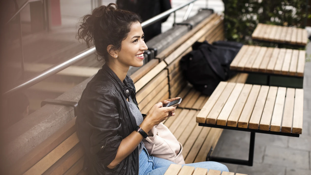 Woman wearing a black leather jacket sitting on a wooden bench with mobile phone on hand  