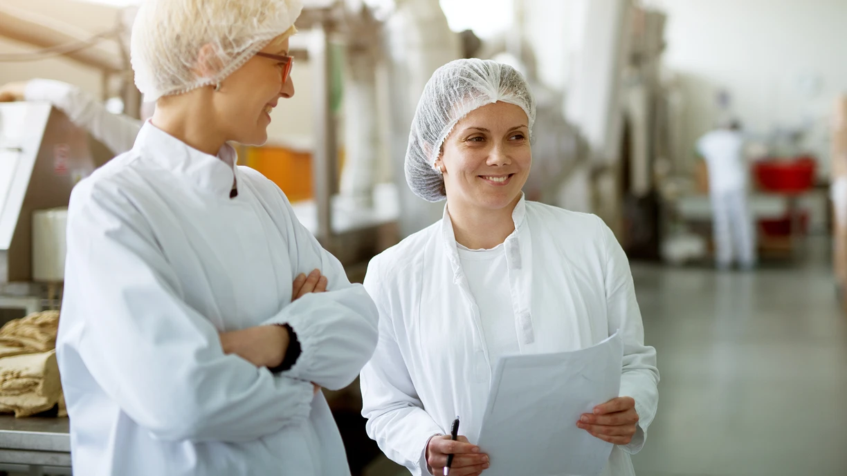 Two female workers in a cheese plant having a chat while one of them holds papers with data 