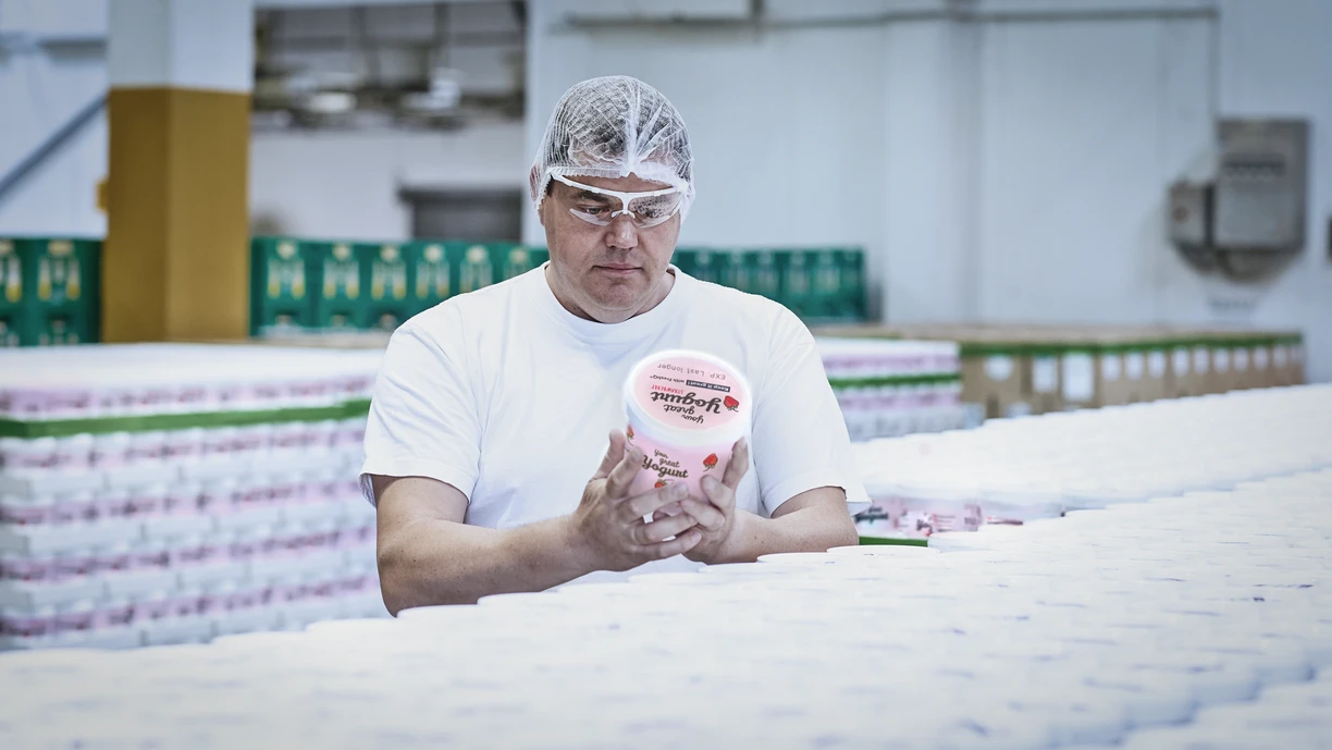 A worker in a dairy in a hairnet and protective goggles examining a pot of fruit yogurt 