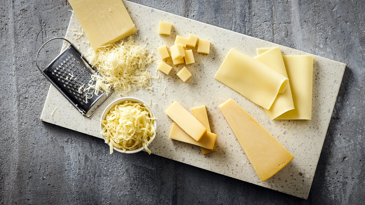 A variety of cheese types including shredded, sliced, cubed, and block pieces arranged on a speckled cutting board with a metal grater and a bowl of shredded cheese, set on a gray textured surface. 