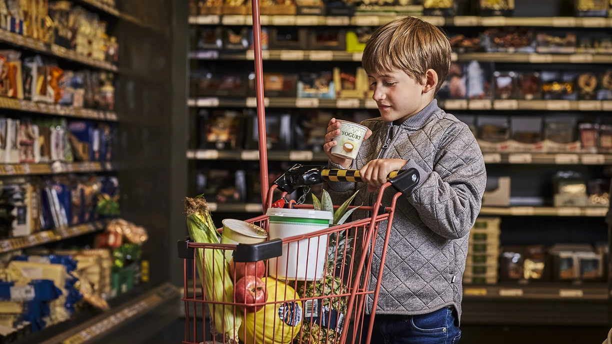 Little boy in a supermarket pushing a child-sized shopping cart and reading the text on the lid of a yogurt pot 