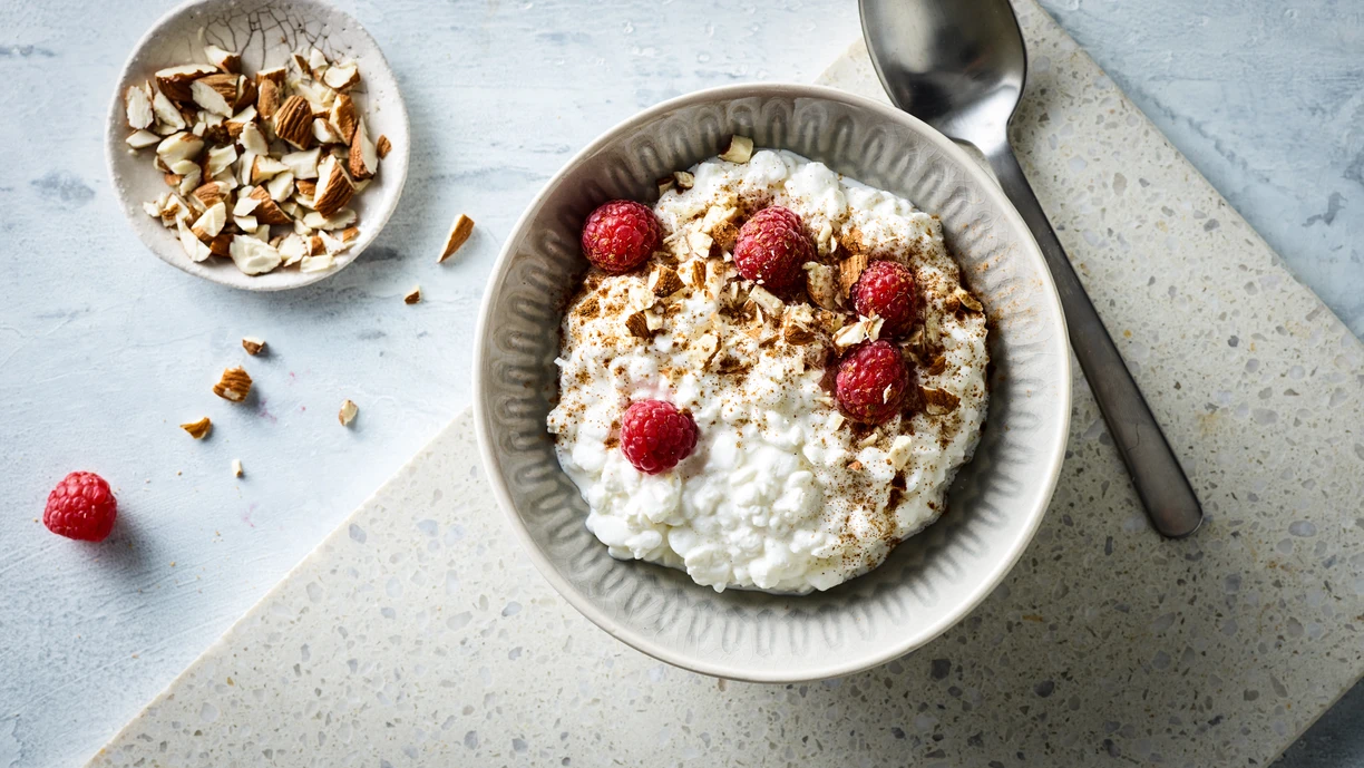A bowl of sweetened cottage cheese with cinnamon, raspberries and chopped almonds. There is a tablespoon beside it, and a bowl of chopped almond and one raspberry on the table behind it. 