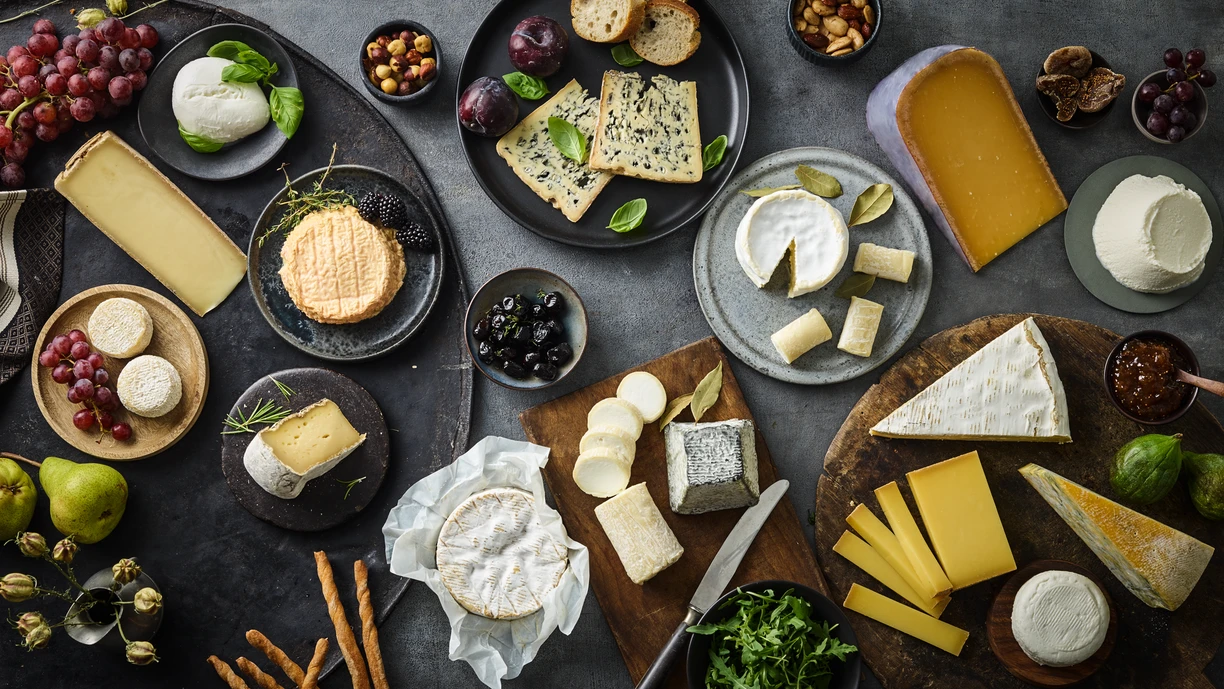 Table spread with three cheeseboards with Brie, goats cheese, Comte, blue cheese, Camembert and various other cheeses. There are also crackers, breadsticks, fruit and nuts on the table. 