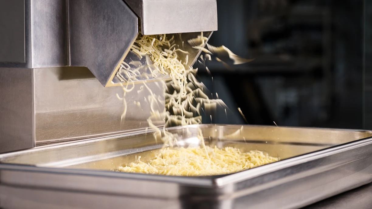 Close-up of a machine shredding cheese into a metal tray, with shredded cheese falling from the chute and forming a pile below.