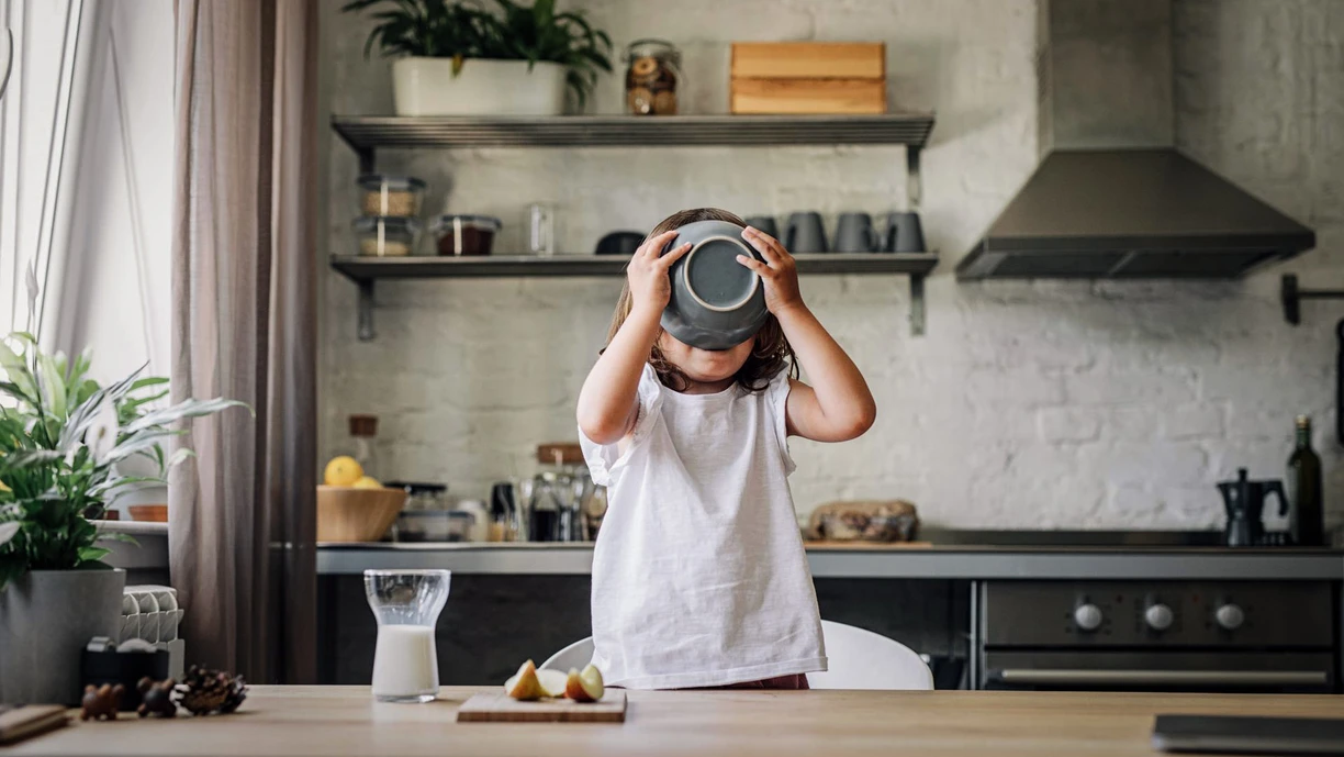 A little boy standing on a chair to reach a kitchen counter, drinking from a bowl of yogurt so that his face is covered by the bowl 