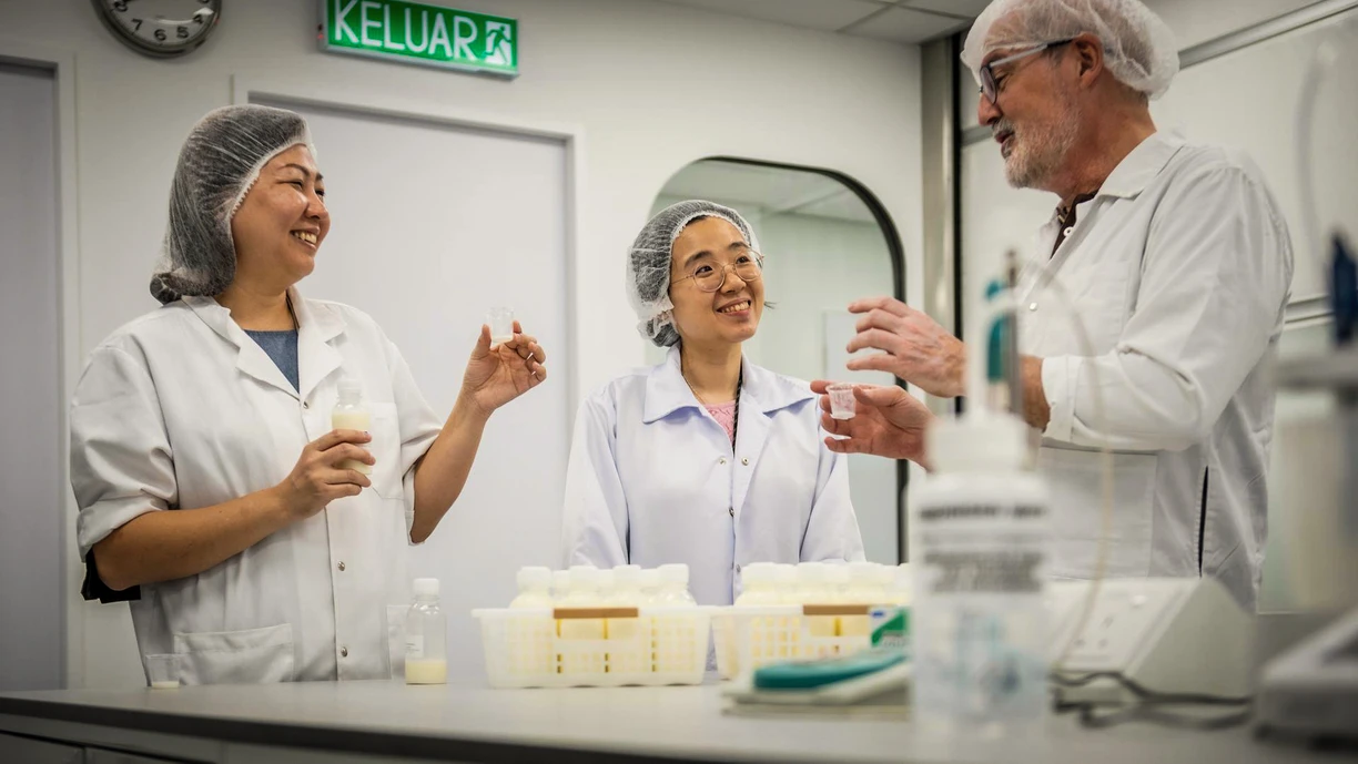 Two female and one male worker in a dairy lab, laughing together as they try samples. All are wearing lab coats and hairnets.
