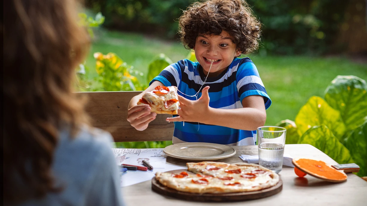 Young boy eating pizza. A cheese string is running from his mouth to the slice and he’s excited by the cheese stretchability. 