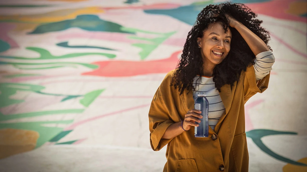 A woman holding a bottle of drinkable yogurt as she pushes back her hair. She’s in an outside setting with colorful patterns painted on the ground in the background. A woman holding a bottle of drinkable yogurt as she pushes back her hair. She’s in an outside setting with colorful patterns painted on the ground in the background. 