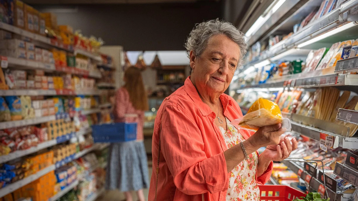 Elderly woman looking at the label on a wedge of plastic-wrapped cheese in the dairy aisle of the supermarket 