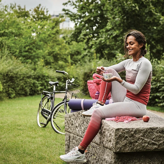 A young woman on a park bench wearing workout clothes with a bicycle in the background, eating from a pot of spoonable yogurt 