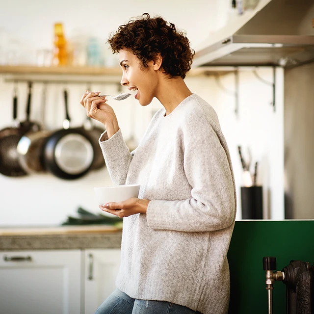 A young woman in her kitchen eating from a pot of spoonable yogurt 