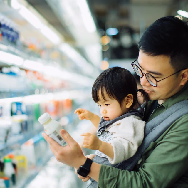 A young father with his daughter in front-facing baby carrier on his chest. Both are looking at the label of a drinkable yogurt.