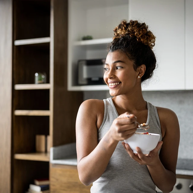 A woman in her kitchen eating from a bowl of spoonable yogurt, granola and berries 
