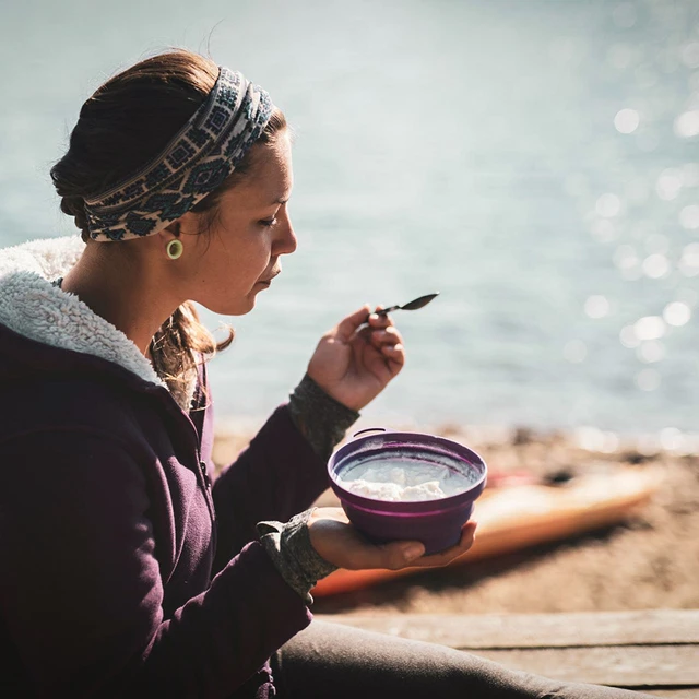 A young woman sitting by the sea with kayaks in the background, eating from a pot of spoonable yogurt 