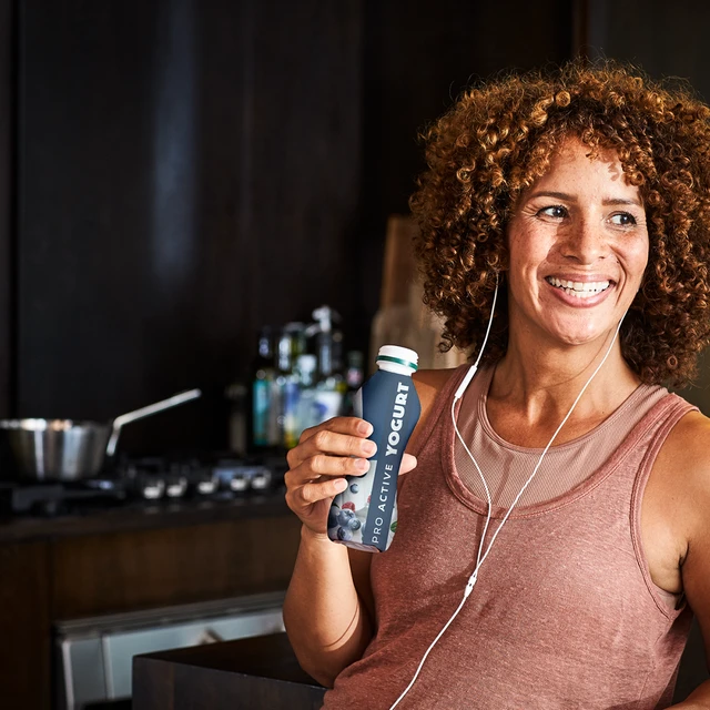 A woman in workout clothes in a café drinking from a bottle of drinkable yogurt with the label “pro active yogurt” v