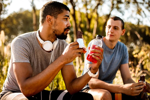 Two men outside in nature, wearing running clothes. One is eating a protein bar and the other
