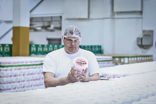 Male worker in a dair wearing a hairnet and white clothes, examining the label on a pot of strawberry yogurt