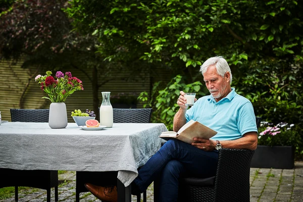 Man sitting outdoors at a table holding a cup and an open book. The table has a vase of flowers, a plate of food, and a bottle of milk. Greenery and trees are in the background