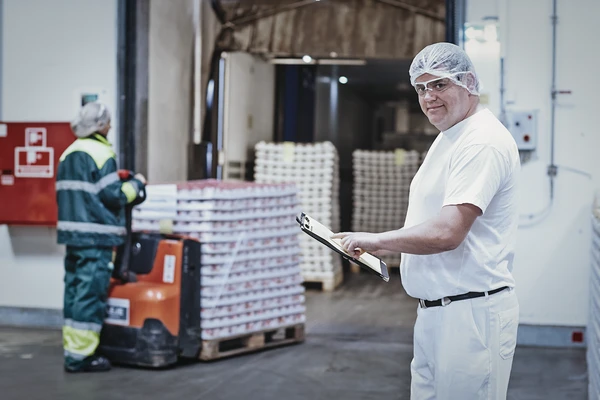 Male dairy worker in hairnet and white clothes, standing outside a storage facility holding a clipboard. In the background another worker is moving a pallet of yogurt tubs with a pallet jack. 