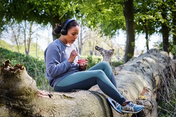 Woman eating a pot of spoonable fruit yogurt while sitting on a fallen tree in a park and wearing running shoes and leggings 