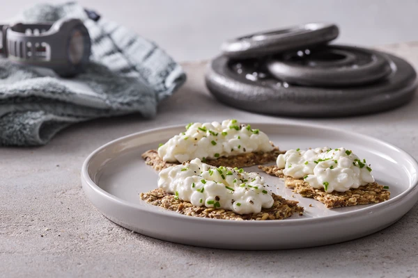 Three high-fiber crackers spread with cottage cheese, with weights and a fitness tracking watch in the background 