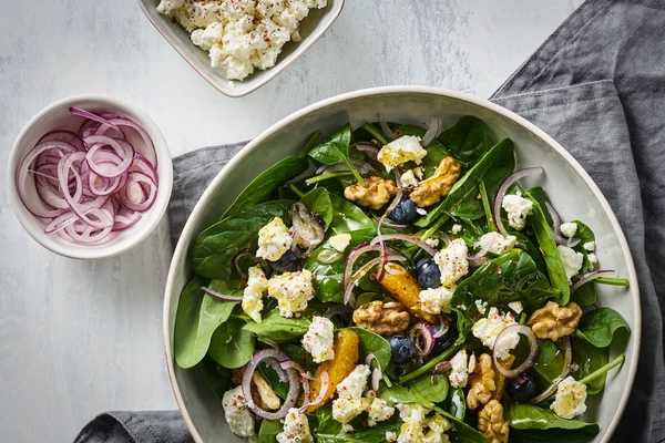 Fresh salad in a large bowl with mixed greens, walnuts, blueberries, orange slices, red onion rings, and crumbled feta cheese. The bowl sits on a light surface with a gray cloth nearby, and two smaller bowls in the corner hold pickled red onions and more crumbled feta cheese.