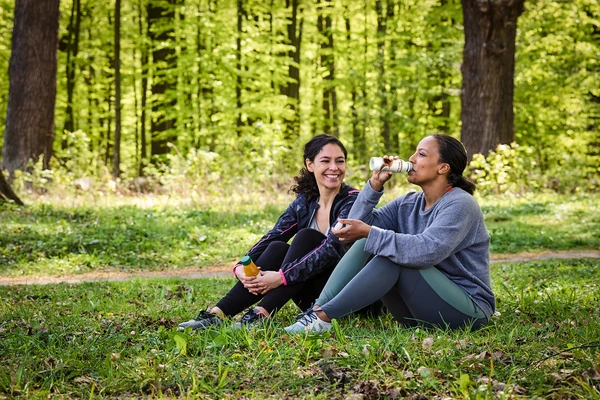 Two women in workout clothes, sitting on the ground in a forest. One is laughing and the other is smiling as she drinks from a bottle of drinkable yogurt 