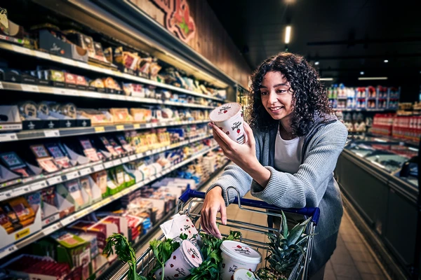 A young woman in the dairy aisle of a supermarket looking at the label on a pot of strawberry yogurt 