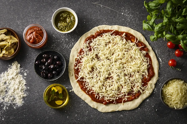 Uncooked pizza with various ingredients in small bowls, including mozzarella 