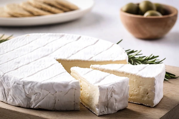 A wheel of Brie with two wedges cut from it and a sprig of rosemary. In the background is a bowl of olives