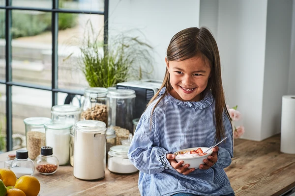 Little girl sitting on a wooden kitchen counter, smiling as she holds a bowl of spoonable yogurt, granola and chopped strawberries 
