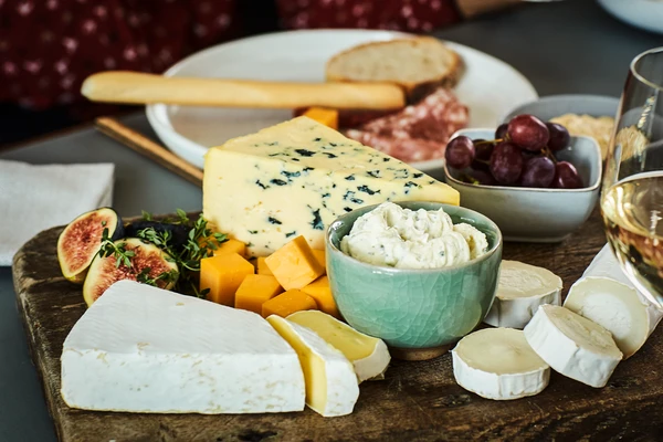 A wooden cheese board with blue cheese, brie, cheddar and goats cheese on a table with people are drinking red wine 
