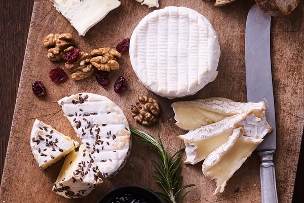 A range of soft cheeses on a wooden board with a sprig of rosemary and some walnuts. 