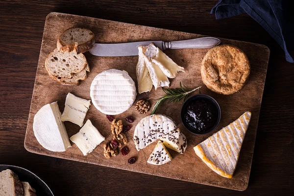 A range of soft cheeses on a wooden board, placed on a wooden table, with a small bowl of fruit preserve, some walnuts and dried