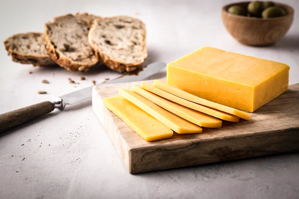 Sliced block of cheddar cheese on a wooden cutting board with multigrain bread, green olives, and a knife. 