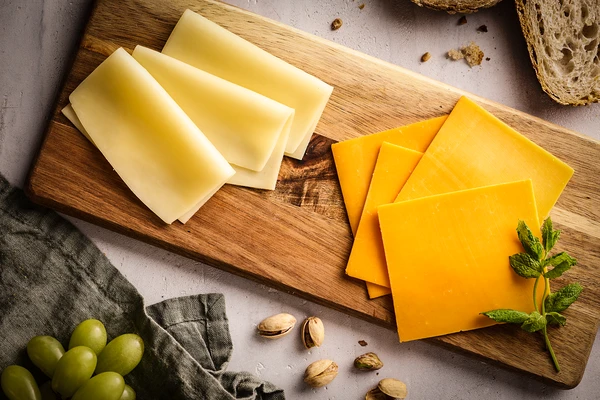 Wooden cutting board with sliced cheddar and Swiss cheese, whole grain bread, pistachios, green grapes, and a mint sprig. 