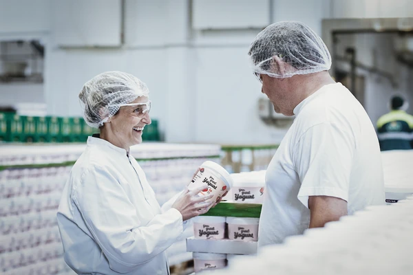 Two dairy workers talking and smiling as one holds a large pot of strawberry yogurt 