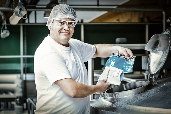 Worker in white uniform and hairnet pouring ingredients into a stainless-steel tank in a food processing facility.