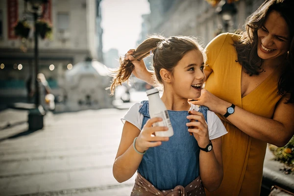 A mother and young daughter laughing together on a sunny street. The daughter is holding a bottle of drinkable yogurt 