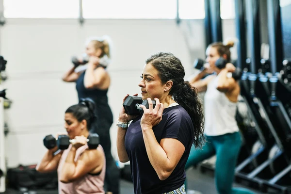 A woman lifting weights at a gym, with other women in the background doing the same. 