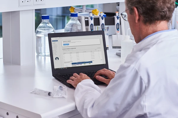 Scientist in a white lab coat, seated in a laboratory, thoughtfully working on a laptop