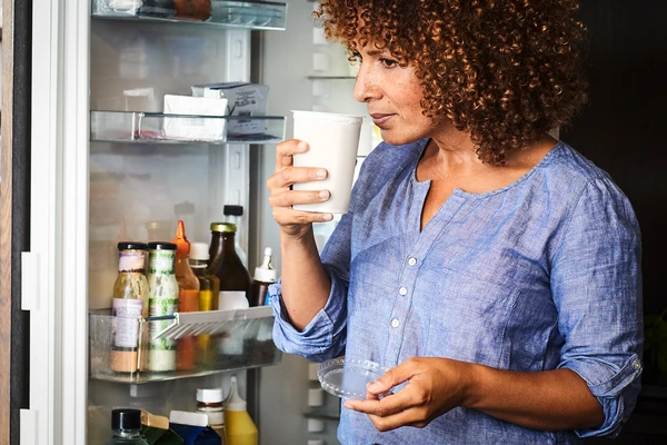 A woman standing by an open fridge, cautiously sniffing an open pot of yogurt to test its freshness 
