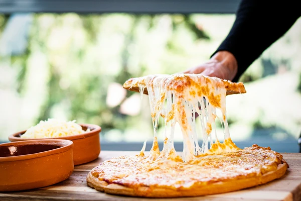 Pizza being sliced on a board. Cheese strings are leading from the slice to the board, showing cheese stretchability. 