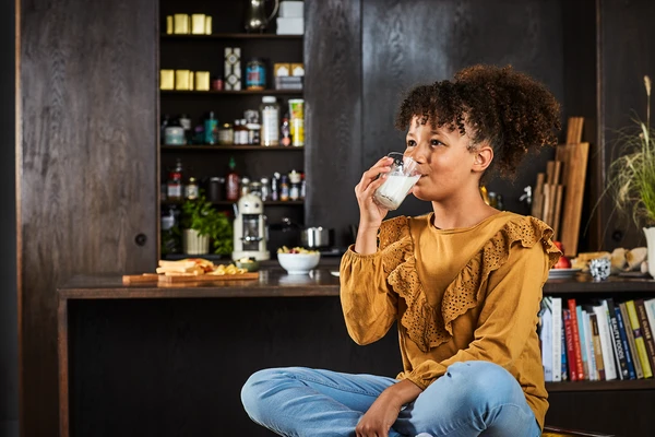 A girl with curly hair sitting cross-legged on a kitchen counter, drinking a glass of milk. She wears a mustard yellow ruffled top and blue jeans