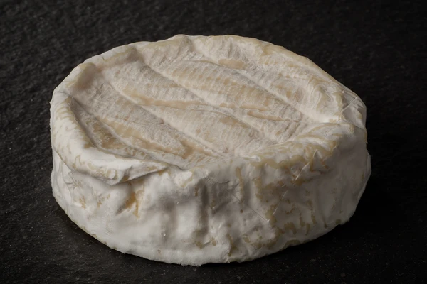 Wheel of Camembert cheese with a white, wrinkled rind on a dark surface—aged to improve cheese flavor and enhance sulfur and garlic notes. 
