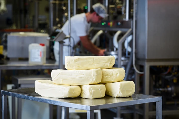 Stack of pasta Filata cheese on a stainless-steel table in a food processing facility, with a worker in white clothing and hairnet operating machinery in the background. Ideal for dairy production and yield optimization. 
