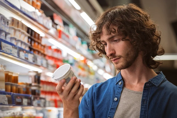 A young man in the dairy aisle of the supermarket looking at the label of a pot of yogurt 